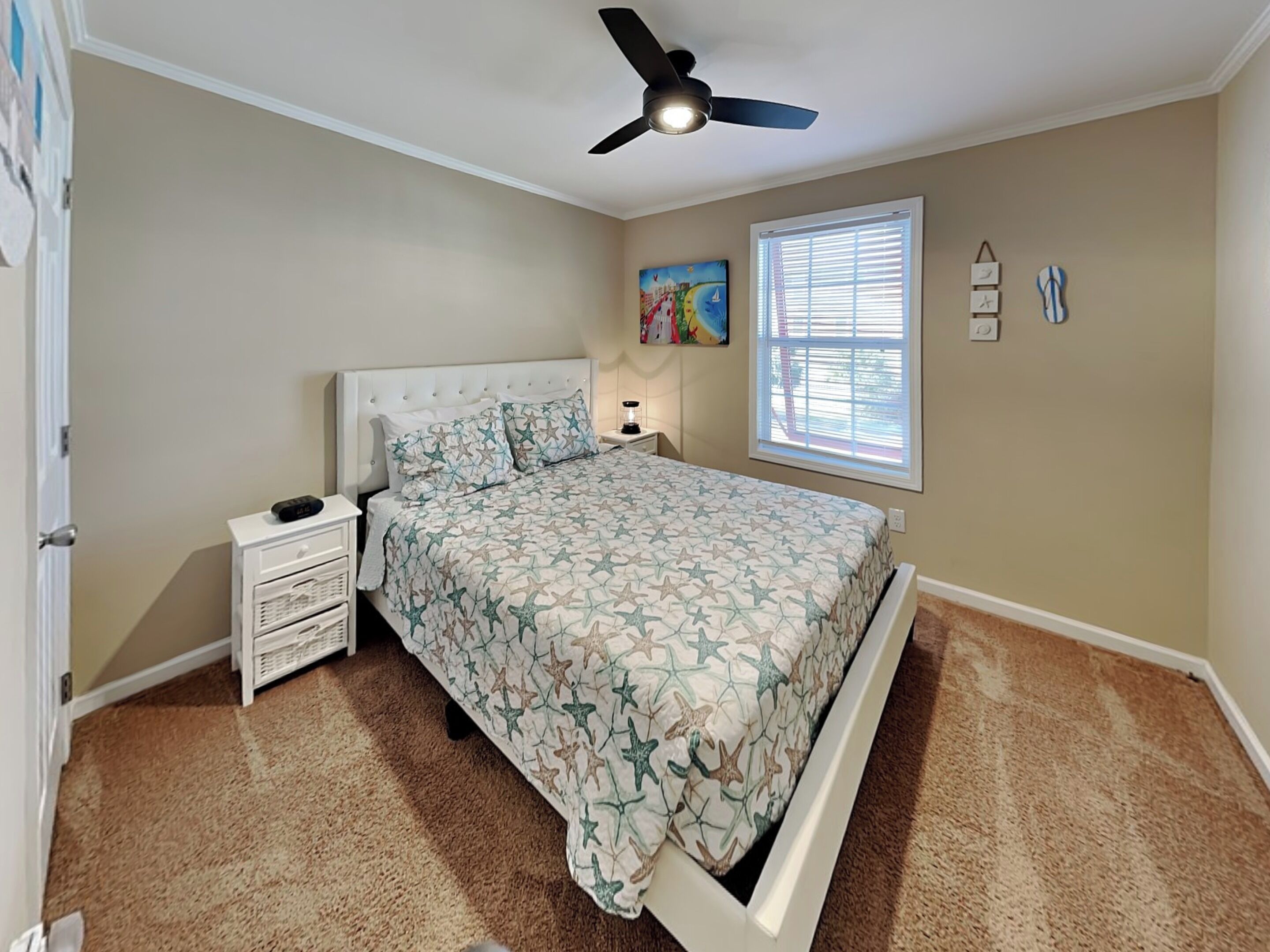 Une chambre à coucher comprenant un lit, des tables de chevet, un ventilateur de plafond, une fenêtre avec des stores et une horloge murale.
