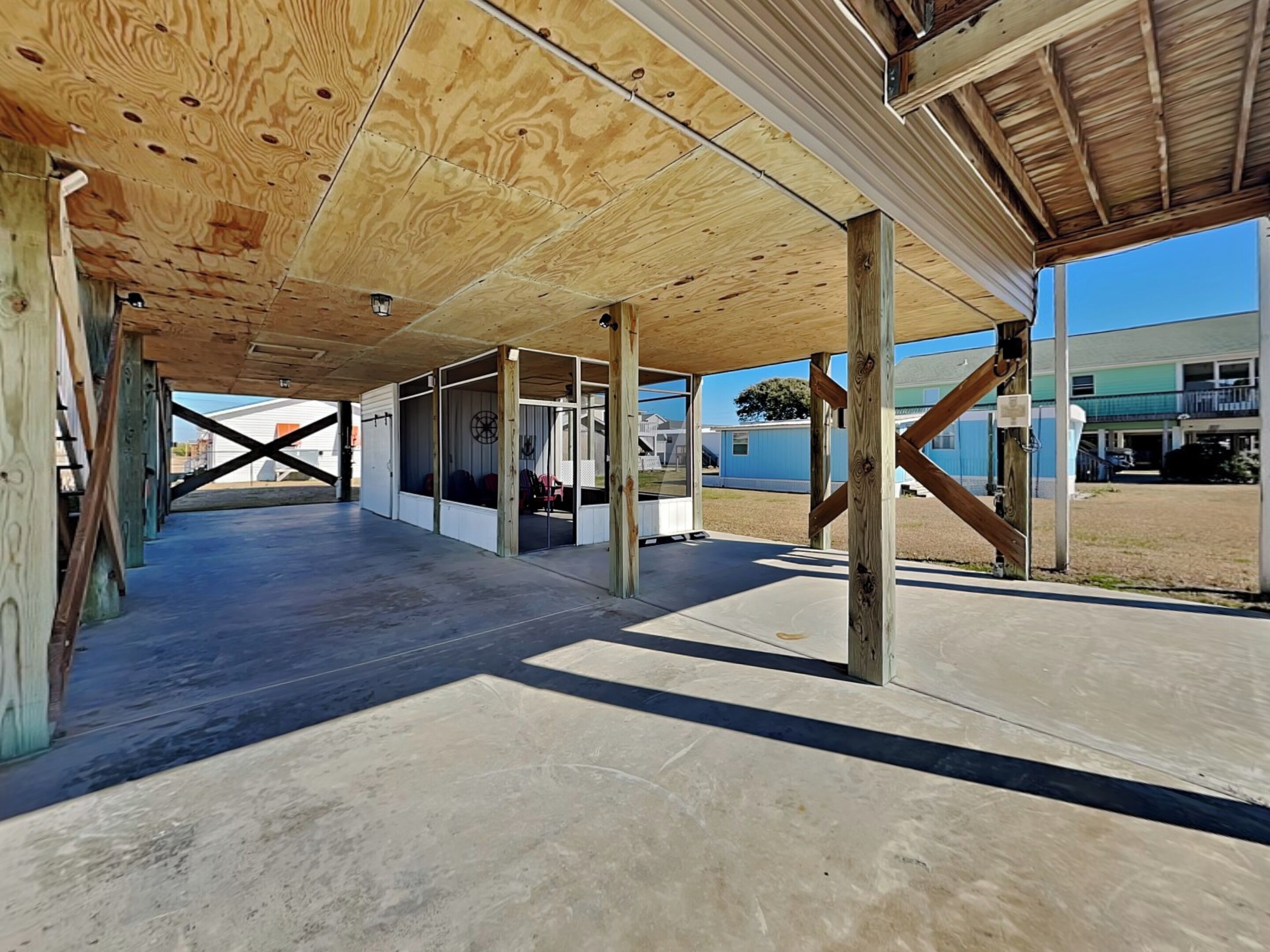 An open, covered outdoor area with wooden beams and concrete flooring, featuring large windows and a view of a building in the background.