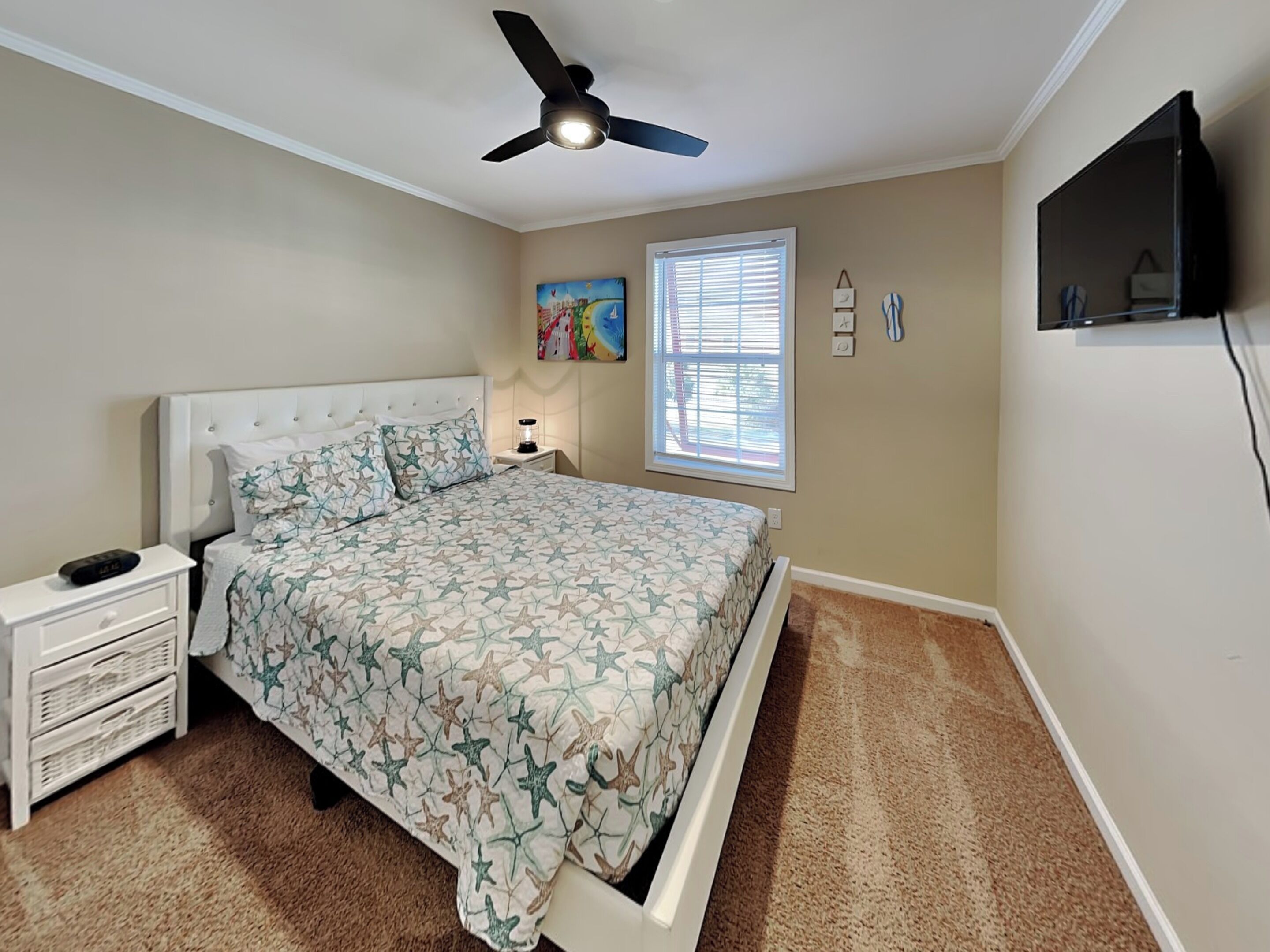 A bedroom with a bed, nightstand, lamp, ceiling fan, window, and wall-mounted TV.
