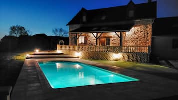 A swimming pool with a stone wall and wooden deck, surrounded by a house with a stone facade and a balcony.