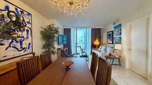 A dining area with a wooden table, chairs, a chandelier, a large abstract painting, a potted plant, and a view of the outdoors. - Urban One Retreat (Miami)