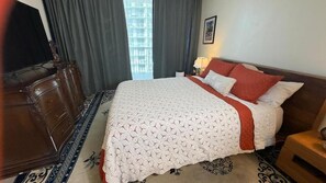Una habitación de hotel con una cama grande, un aparador de madera, una mesita de noche con una lámpara y vistas a la ciudad por la ventana.