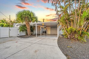 A covered outdoor area with a concrete floor, surrounded by palm trees and a white fence. - Key Life Sands | Duplex Unit Near Crescent Beach with Shared Pool (Sarasota)
