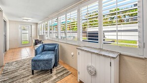 Une pièce avec une chaise bleue, un meuble blanc et une grande fenêtre avec des stores.