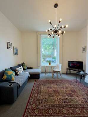 A living room with a sofa, a TV, a chandelier, and a large window. - Stylish Apartment Near Town Centre (Gloucester)