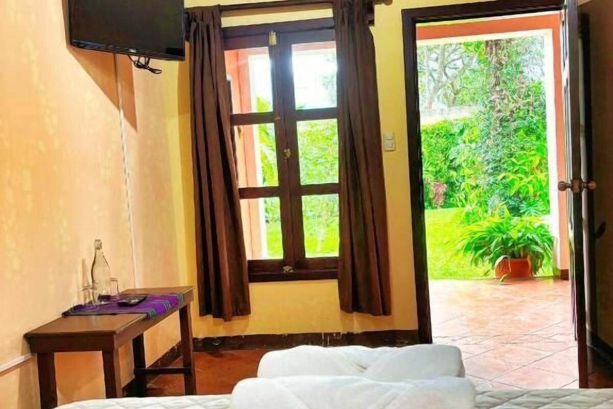 Ein Zimmer mit einem Bett, einem Fernseher, einem Fenster mit Vorhängen und Blick ins Grüne.