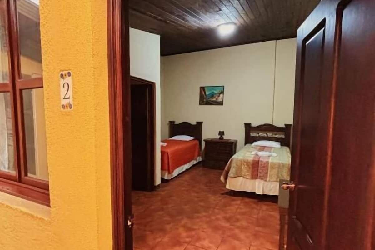 Ein Zimmer mit Fliesenboden, einem Bett mit Steppdecke, einem Nachttisch und einem Bild an der Wand.