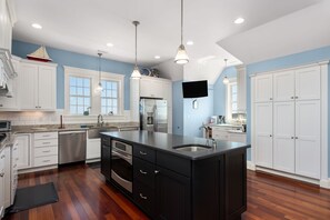 A modern kitchen with white cabinetry, a large island, stainless steel appliances, and pendant lighting. - Lone Cedar II: Soundfront, Amazing Sunset Views, Private Pool (Nags Head)