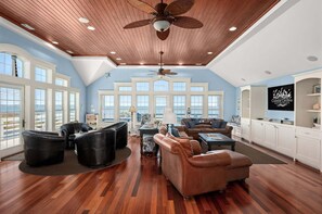 A spacious living room with a high ceiling, large windows, and a view of the ocean. - Lone Cedar II: Soundfront, Amazing Sunset Views, Private Pool (Nags Head)