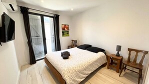 Een slaapkamer met een bed, een aan de muur bevestigde televisie, een stoel, een bureau met een lamp en een raam met gordijnen.