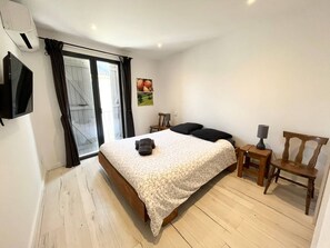 Ein Schlafzimmer mit einem Bett, einem an der Wand befestigten Fernseher, einem Sessel, einem Schreibtisch mit Lampe und einem Fenster mit Vorhängen.
