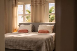 A bed with a quilted cover, two pillows, and a view of palm trees through a window with curtains. - Caribbean Club 111 Parrots Perch on a small-scale resort with beautiful views (Kralendijk)