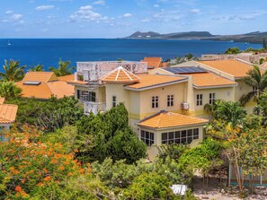 Un quartier résidentiel en bord de mer, avec des maisons dotées de toits en tuiles orange et de panneaux solaires, entouré d’une végétation luxuriante et de palmiers.