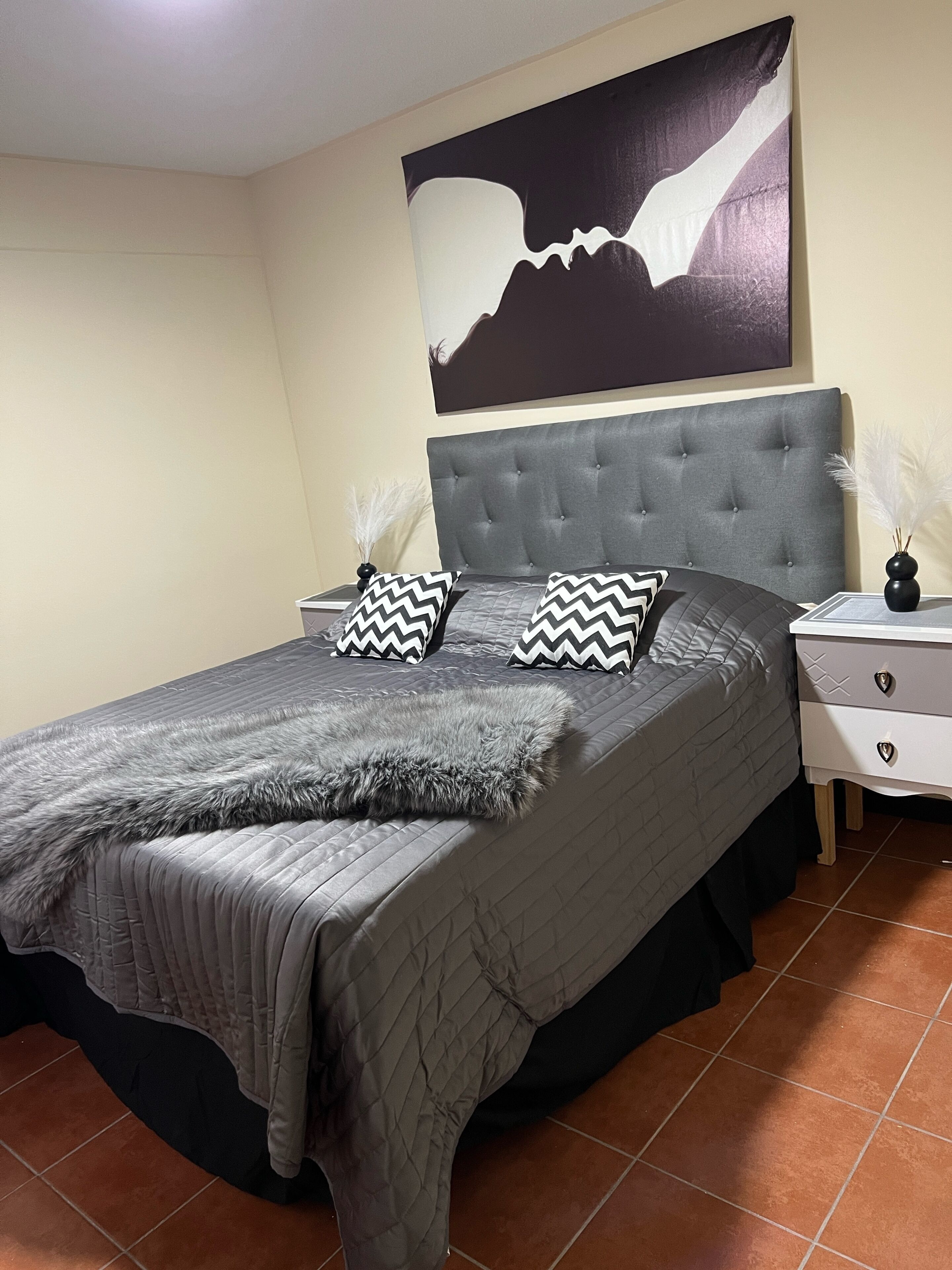 Un dormitorio con un cabecero tapizado en gris, un edredón acolchado en gris, dos almohadas a cuadros blanco y negro, una manta mullida gris y una mesita de noche con un florero de plumas blancas.