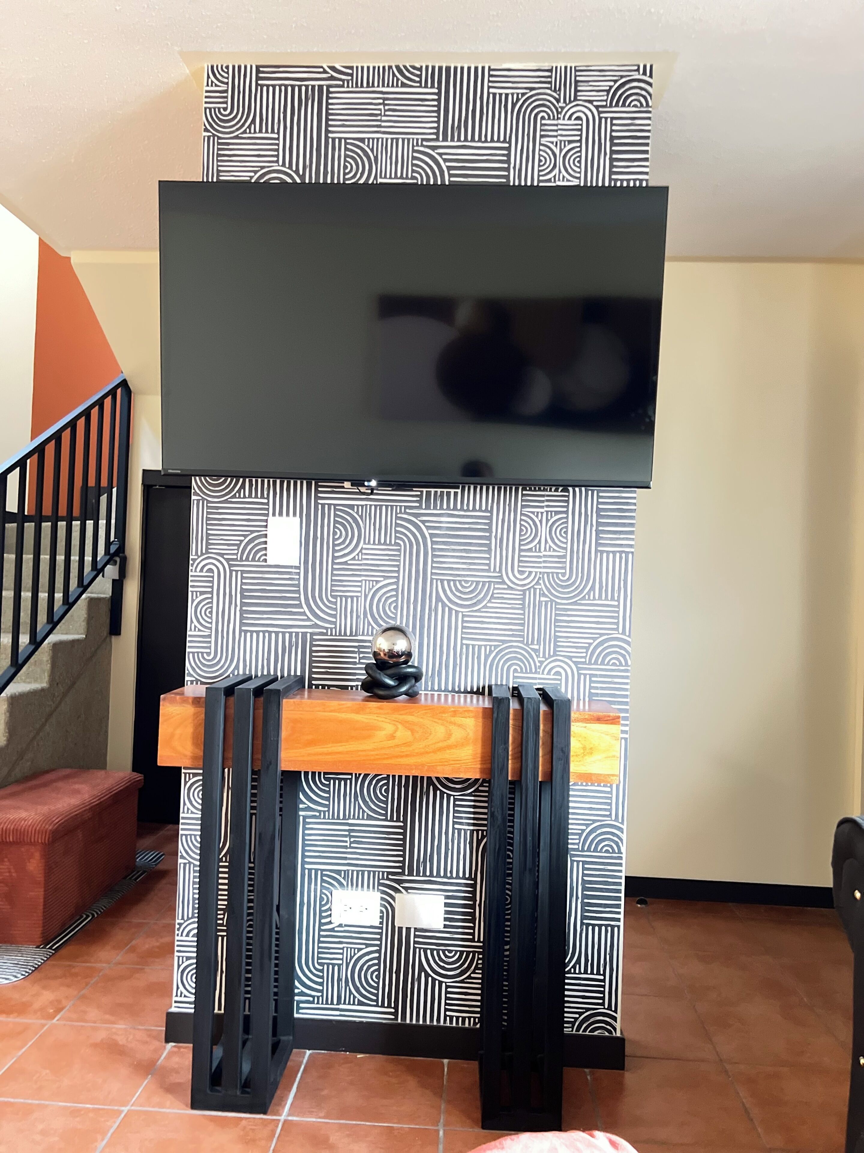 A room with a black and white patterned wall, a flat-screen TV mounted above a wooden desk, and a staircase with a black railing.