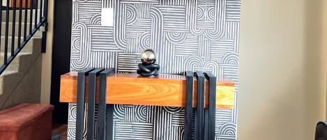 A room with a black and white patterned wall, a flat-screen TV mounted above a wooden desk, and a staircase with a black railing.