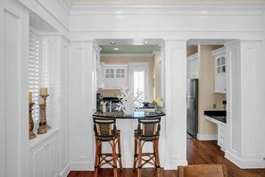 A kitchen with white cabinetry, a central island with two wicker chairs, and a view into the dining area. - Casa Del Mare Caribe (Miramar Beach)