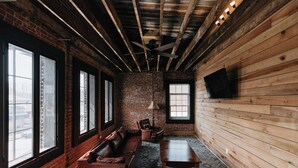 A room with a brick wall, wooden beams, a leather sofa, a wooden table, and a TV mounted on the wall. - Historic Loft Stay Near Mammoth Cave National Park (Cave City)