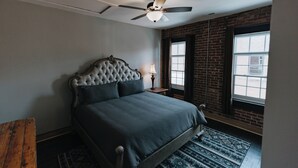 A bedroom with a tufted headboard, a bed, a nightstand with a lamp, a window with a view, and a ceiling fan. - Historic Loft Stay Near Mammoth Cave National Park (Cave City)