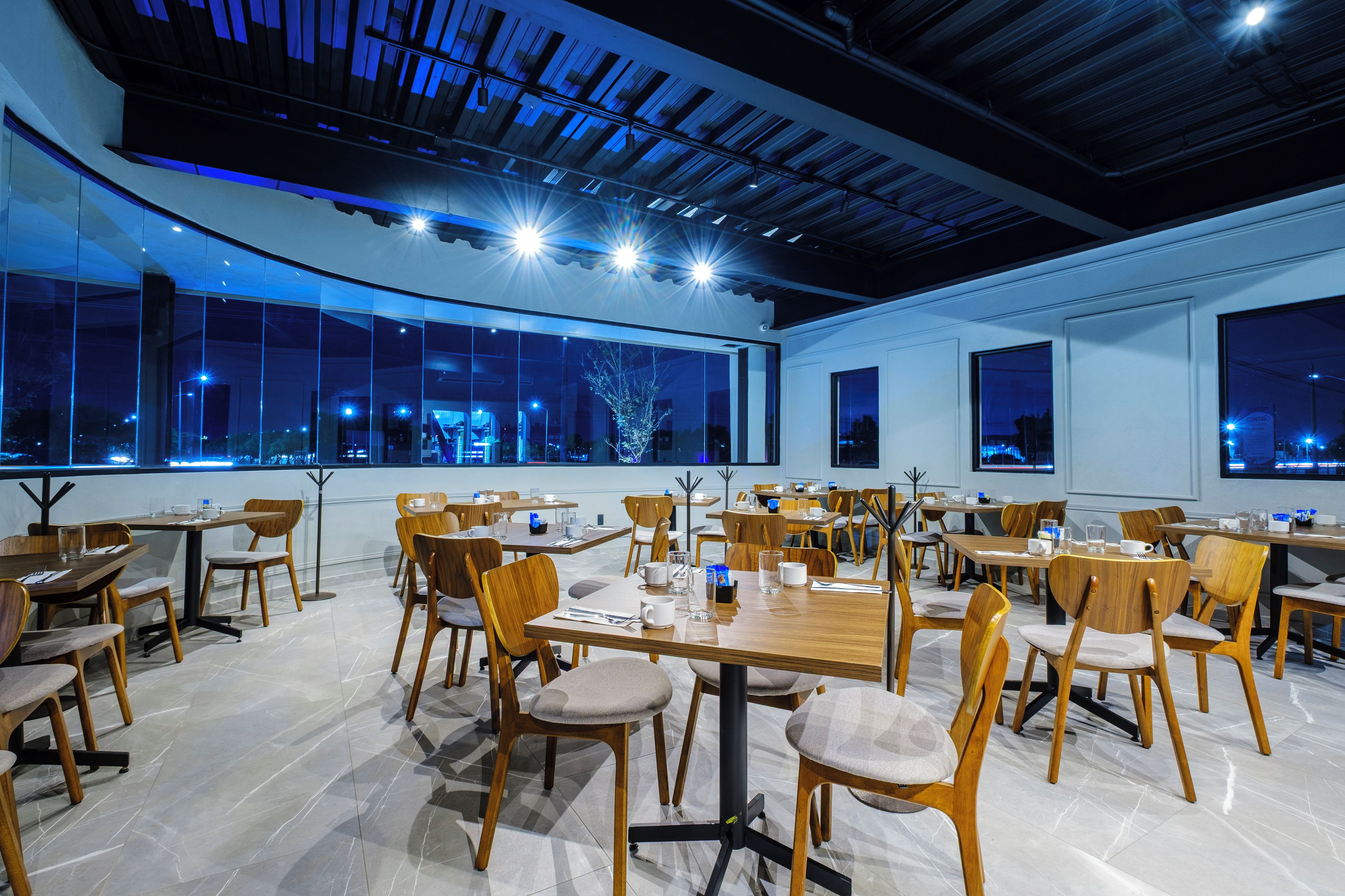 Un comedor moderno con varias mesas y sillas, amplias ventanas que ofrecen vistas a la ciudad y un techo alto con vigas a la vista.