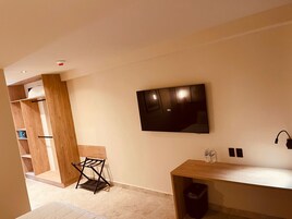 Una habitación moderna con un televisor de pantalla plana montado en la pared, un escritorio de madera con un cesto de basura y una pequeña zona de estar con una silla.
