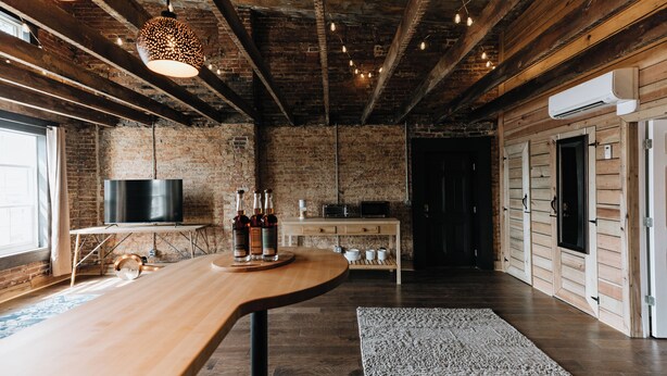 A rustic kitchen with exposed brick walls, wooden beams, a long wooden table, and hanging pendant lights.