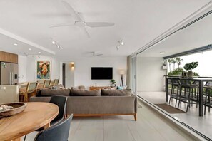 Un salon moderne comprenant un canapé, une table à manger et des chaises, un téléviseur fixé au mur, et un balcon avec des sièges d’extérieur.