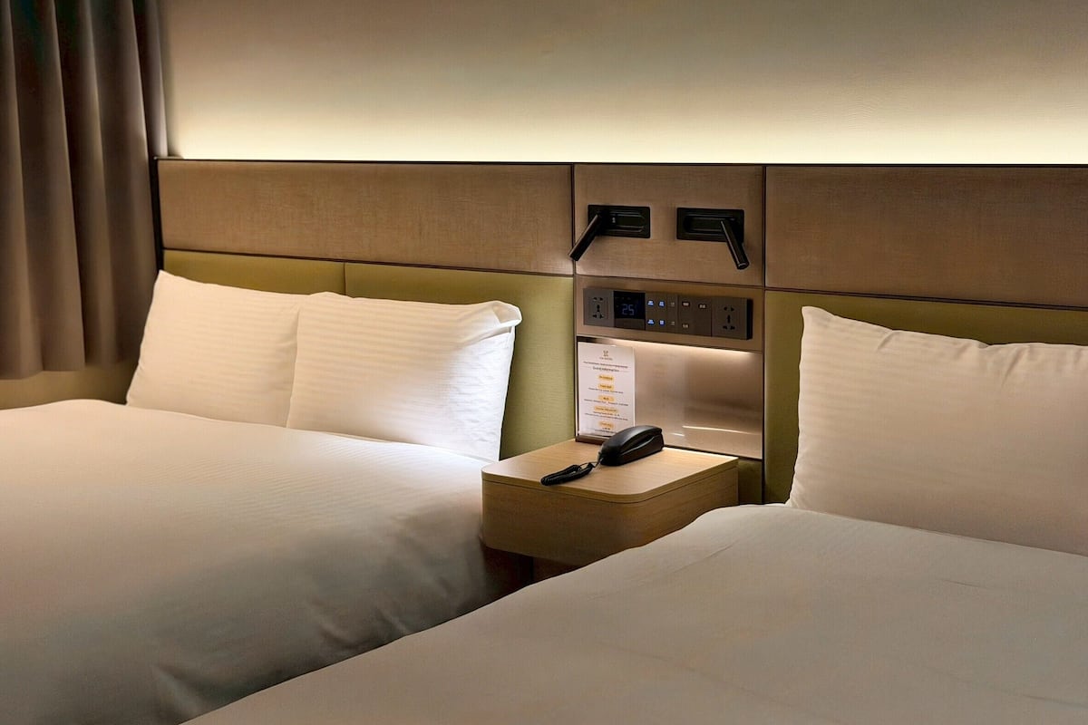 Une chambre d’hôtel avec deux lits, une table de chevet avec un téléphone et un panneau de commande au-dessus des lits.