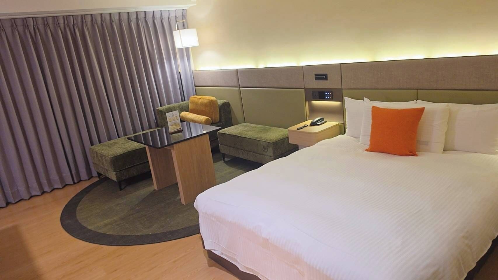 A hotel room with a bed, a desk, a chair, a nightstand, a lamp, and a window with curtains.