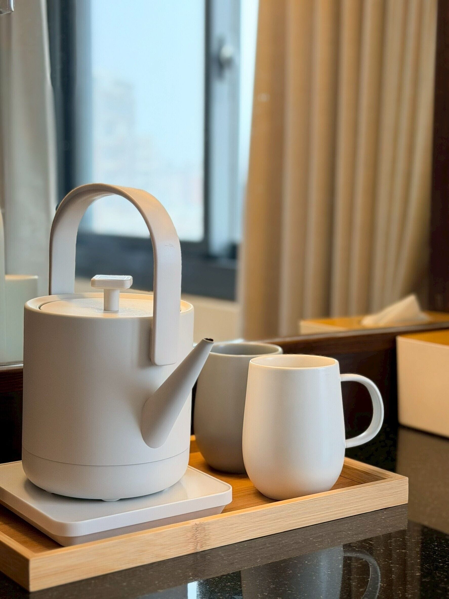 A white teapot and mug on a tray, a window with curtains, and a view of buildings.