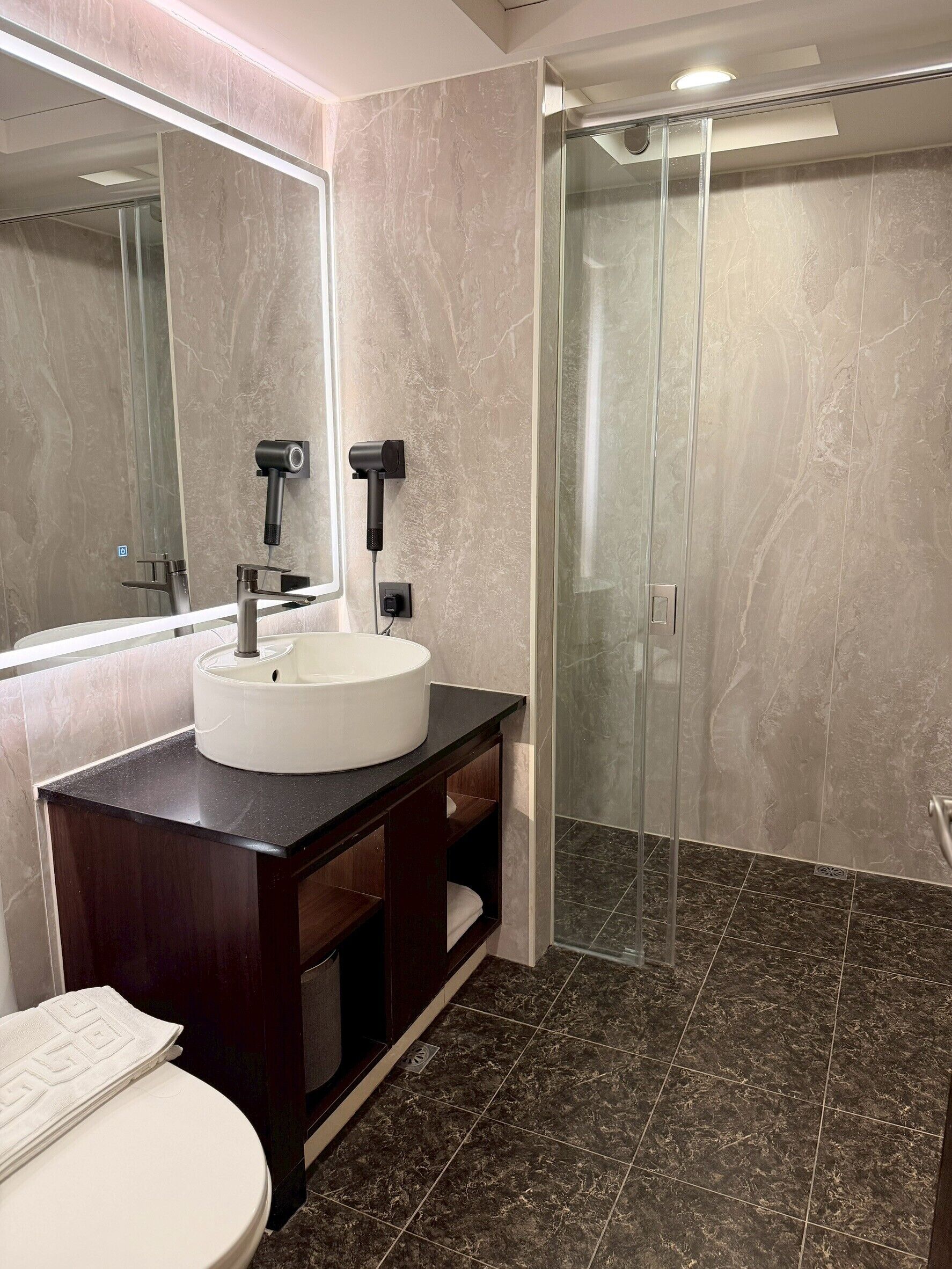 A modern bathroom with a marble vanity, a large mirror, a white sink, a glass-enclosed shower, and a dark tiled floor.
