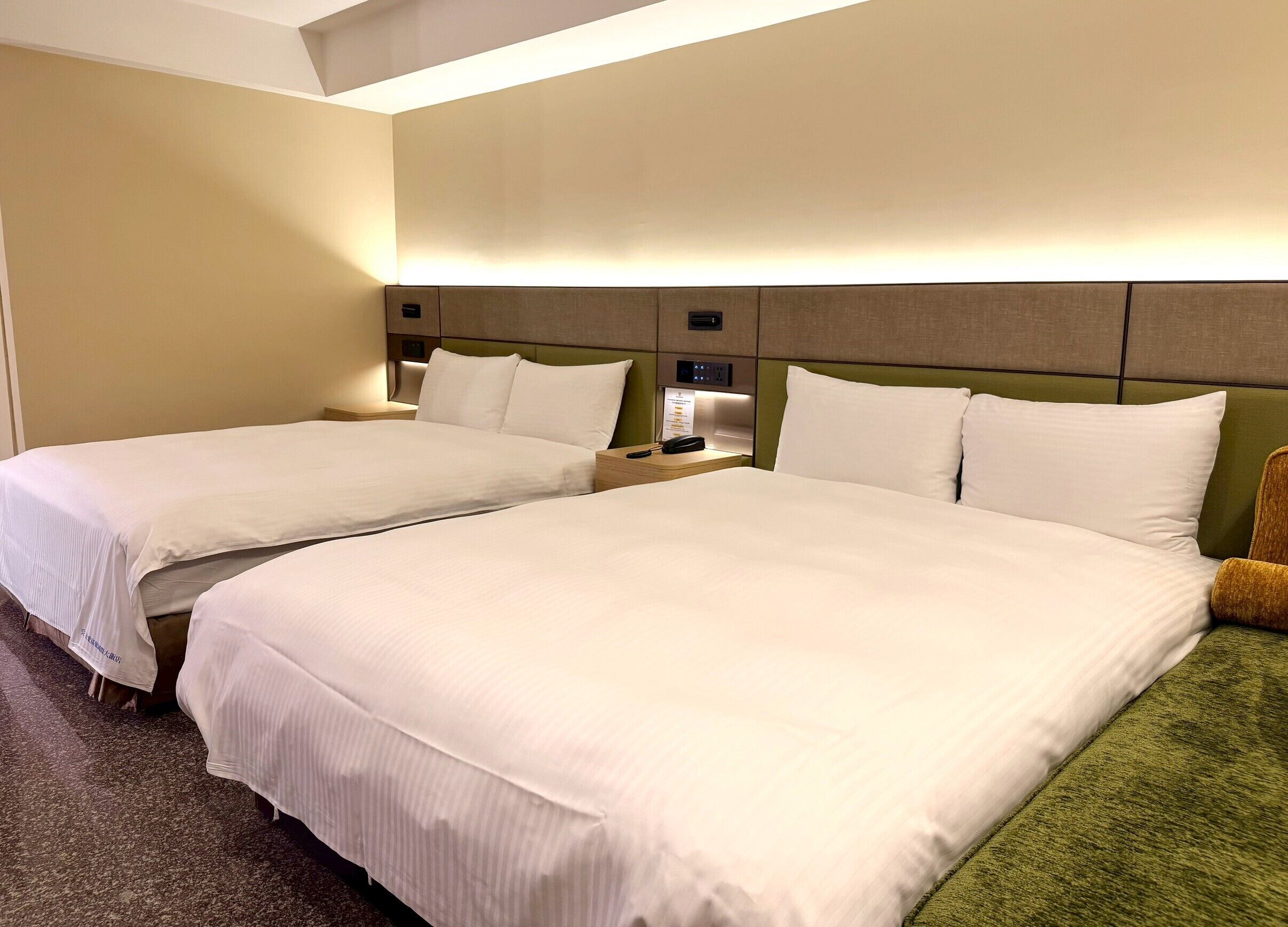 A hotel room with two beds, a nightstand, a lamp, and a green chair.