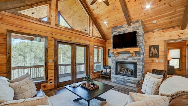 A log cabin interior with a stone fireplace, wooden walls, and large windows. - Riverfront cabin in lovely gated community with hot tub and game room (Ellijay)