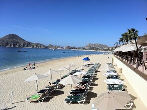 Ein Strand mit Liegestühlen und Sonnenschirmen, einem klaren, blauen Himmel und Booten auf dem Wasser.