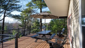 Een houten terras met picknicktafels en een parasol, omgeven door bomen en een huis.