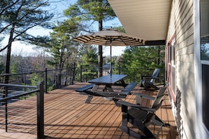 Une terrasse en bois avec des tables de pique-nique et un parasol, entourée d’arbres et d’une maison.