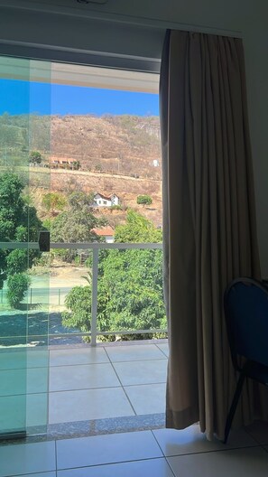 A room with a sliding glass door leading to a balcony with a view of a hilly landscape and a few houses.