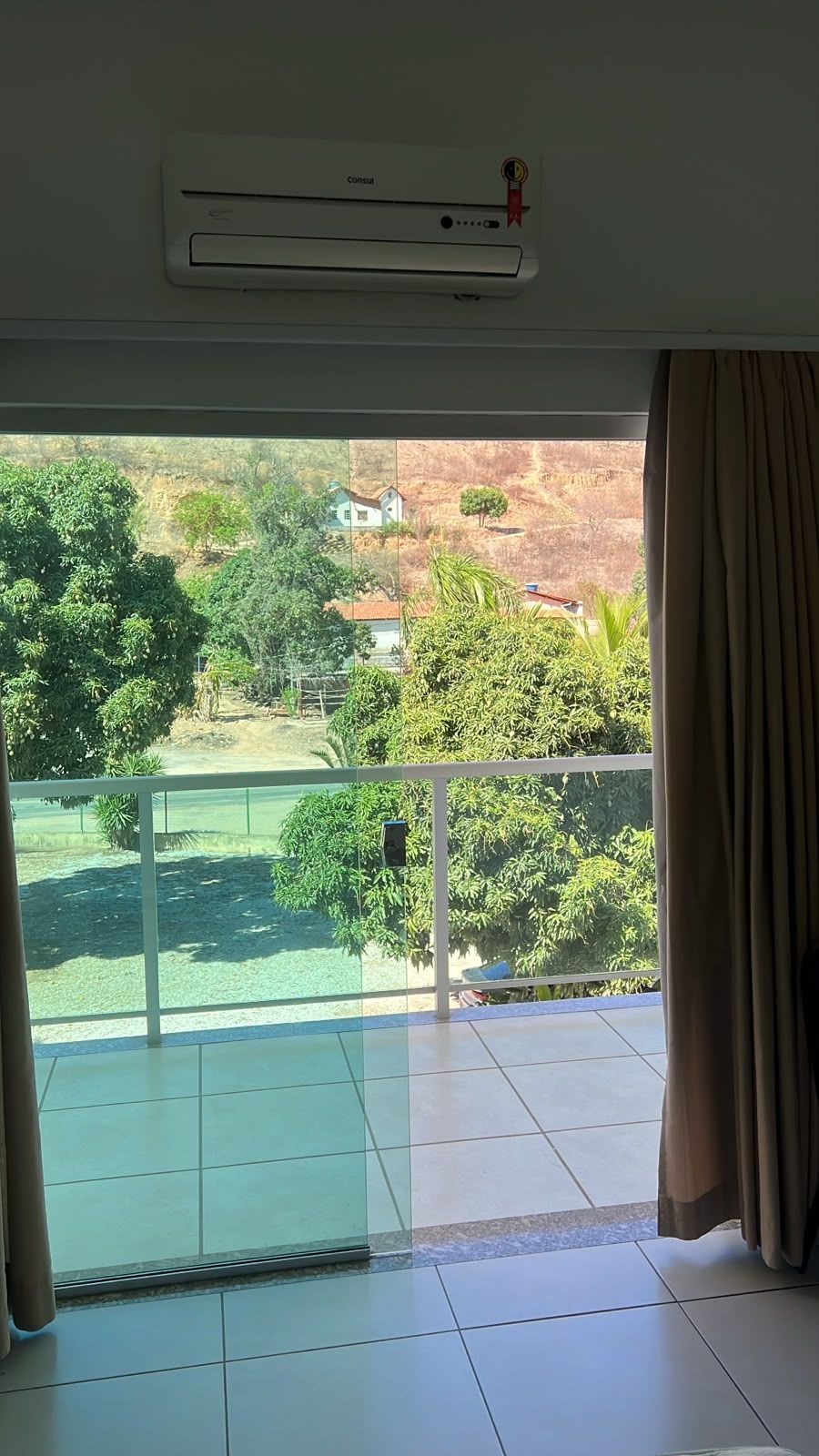 Quarto com vista para uma paisagem verde, varanda com porta de vidro e unidade de ar condicionado na parede.
