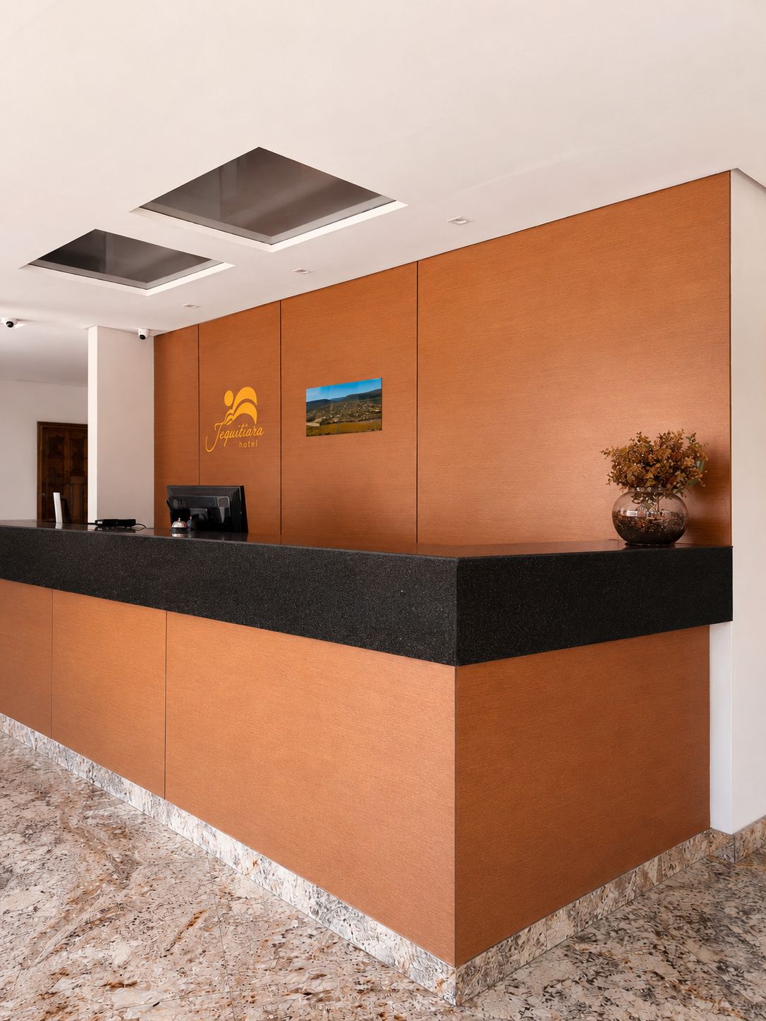 A hotel reception desk with a logo, a coffee machine, and a vase with dried flowers.