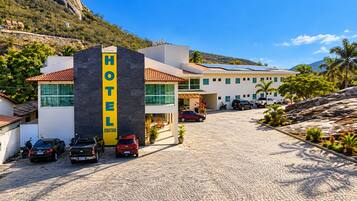 Um hotel com uma placa amarela grande, um acesso pavimentado e montanhas ao fundo.