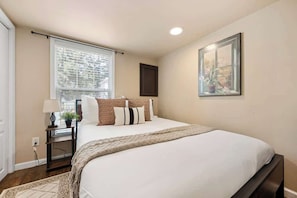 A bedroom with a bed, a nightstand, a lamp, a window with blinds, and a framed picture on the wall. - Woodland Peak Tiny Lodge (Woodland Park)