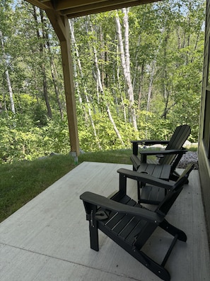 A covered patio with two Adirondack chairs and a table, overlooking a forested area. - RiverWest Trailside Tranquility - Sauna - Dogs Friendly (Duluth)