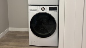 Een wasmachine en droger boven elkaar in een wasserij met een houten vloer en een witte deur.