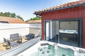 A hot tub with a patio area, a sliding glass door leading to a room with a bed, and a red building exterior. - Cozy Cottage with Spa - Welkeys (Moliets-et-Maa)