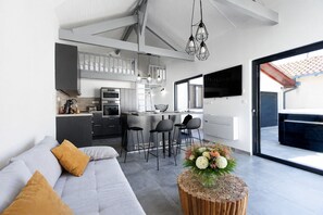 A modern kitchen with a dining area, a sofa with cushions, and a wooden coffee table with flowers. - Cozy Cottage with Spa - Welkeys (Moliets-et-Maa)