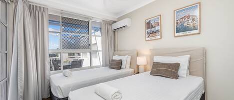 A hotel room with two beds, a ceiling fan, air conditioning, a window with curtains, and two framed pictures on the wall.