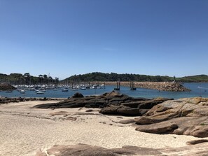 Een strand met een rotsachtige voorgrond, helderblauwe lucht en een jachthaven met boten.
