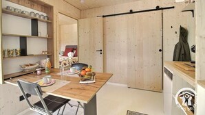 A compact room with a wooden dining table, a chair, a bed with a red pillow, and a coat hanging on the wall. - Ker Lann - TREBEURDEN (TREBEURDEN)