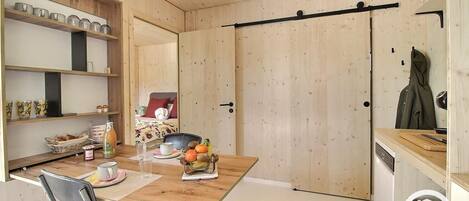 A compact room with a wooden dining table, a chair, a bed with a red pillow, and a coat hanging on the wall.
