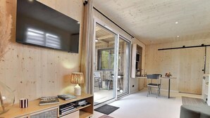 A modern interior with a flat-screen TV, a wooden bookshelf, a dining area with a table and chairs, and a sliding glass door leading to an outdoor space. - Ker Lann - TREBEURDEN (TREBEURDEN)
